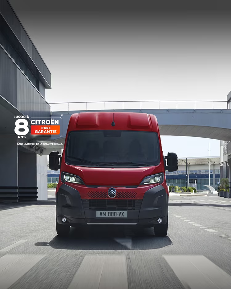 Vue de face d’un fourgon Citroën Jumper rouge roulant dans un environnement urbain moderne, mettant en avant son design robuste, sa praticité et sa présence affirmée sur la route.