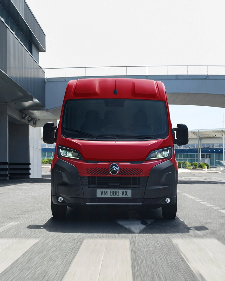 Vue de face d’un fourgon Citroën Jumper rouge roulant dans un environnement urbain moderne, mettant en avant son design robuste, sa praticité et sa présence affirmée sur la route.