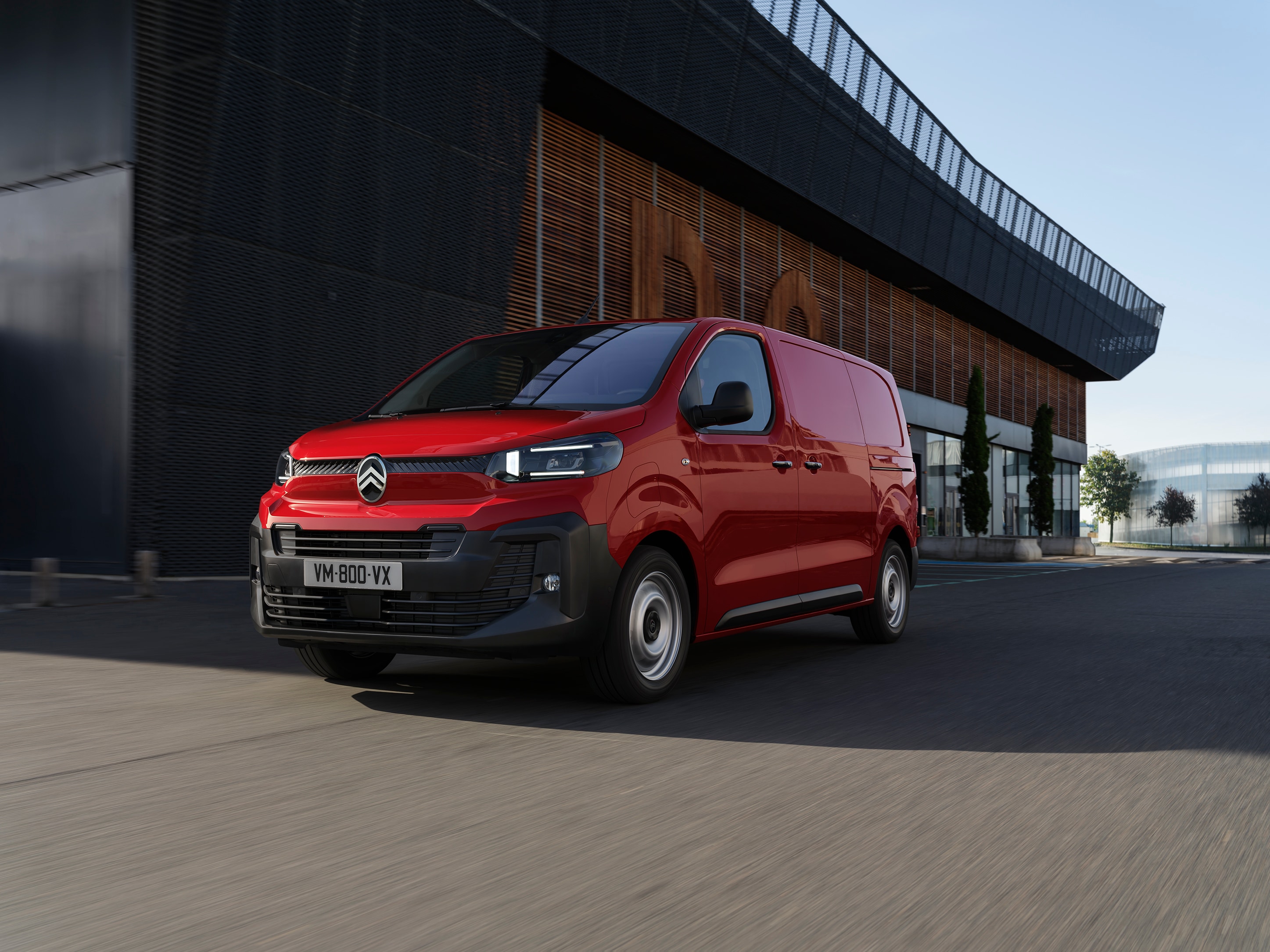 Vue extérieure avant en trois-quarts d’un Citroën Jumpy rouge stationné près d’un bâtiment moderne, illustrant un design professionnel, une grande praticité et une présence assurée.