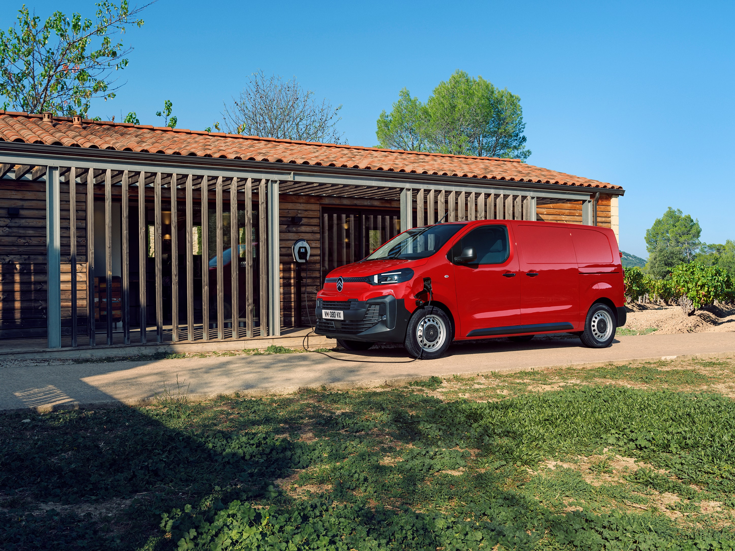 Vue extérieure en trois-quarts latérale d’un Citroën ë-Jumpy rouge en charge devant un bâtiment, illustrant la mobilité électrique, la praticité au quotidien et une présence professionnelle affirmée.