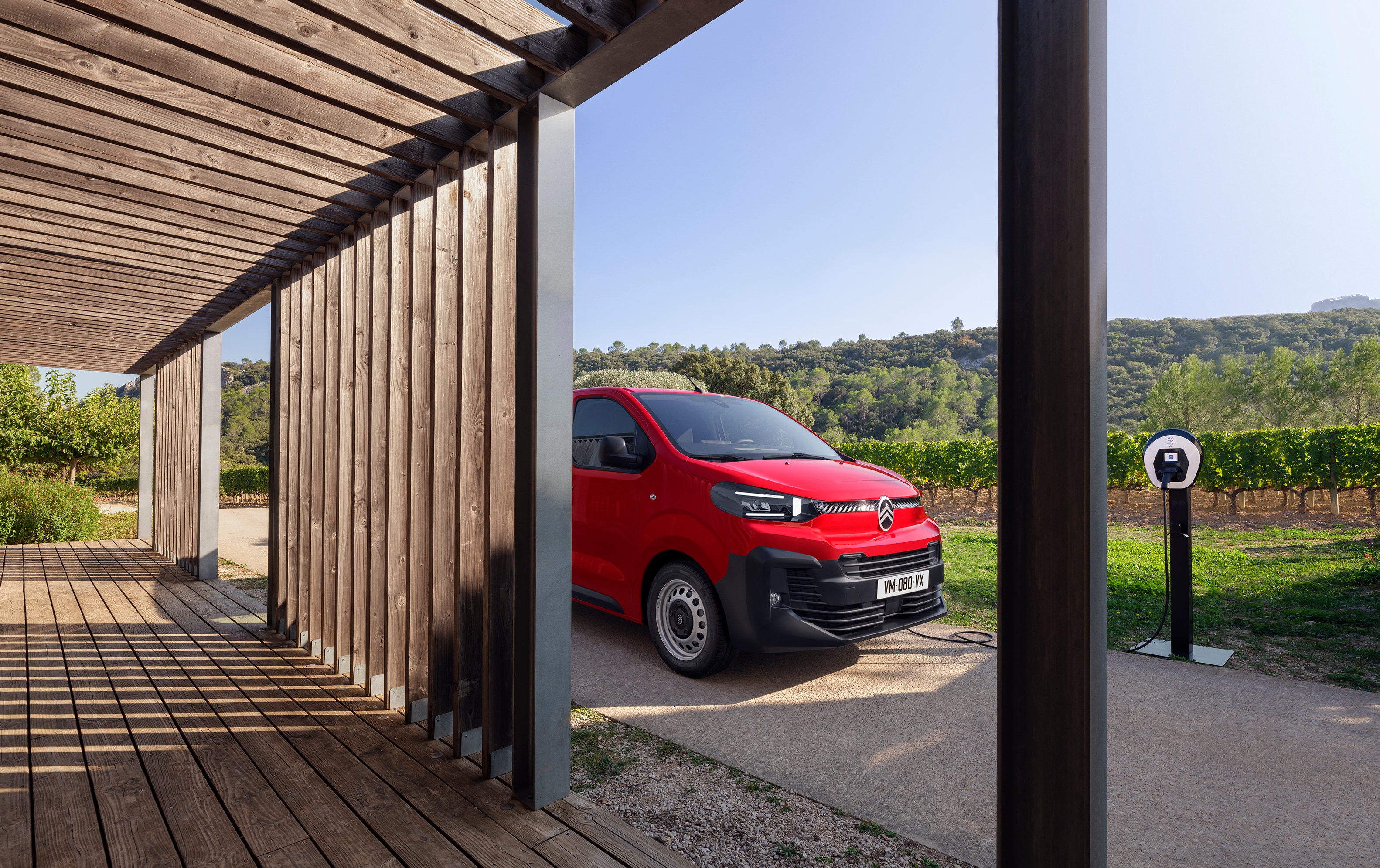 Vue extérieure en trois-quarts latérale d’un Citroën ë-Jumpy rouge stationné devant un bâtiment en bois, mettant en avant un design électrique moderne, la praticité et une image professionnelle.