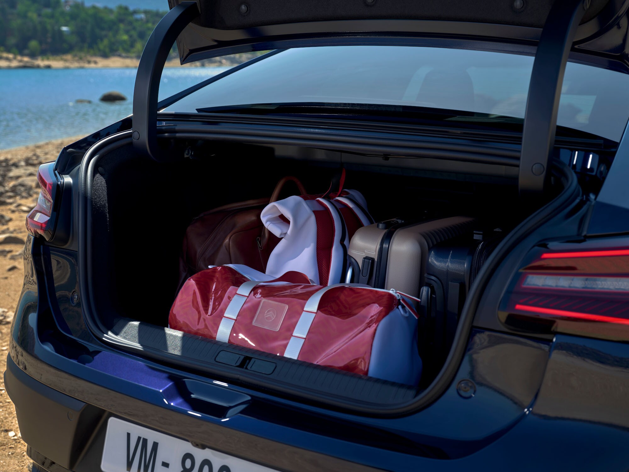 Coffre ouvert de la Citroën ë-C4 X chargé de sacs de voyage à côté d’un emplacement au bord du lac
